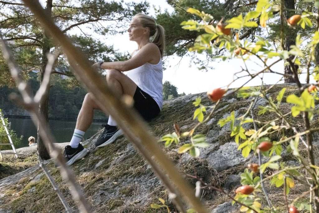 Woman in running kit sitting on a rocky outcrop beside a lake, framed by rosehip branches